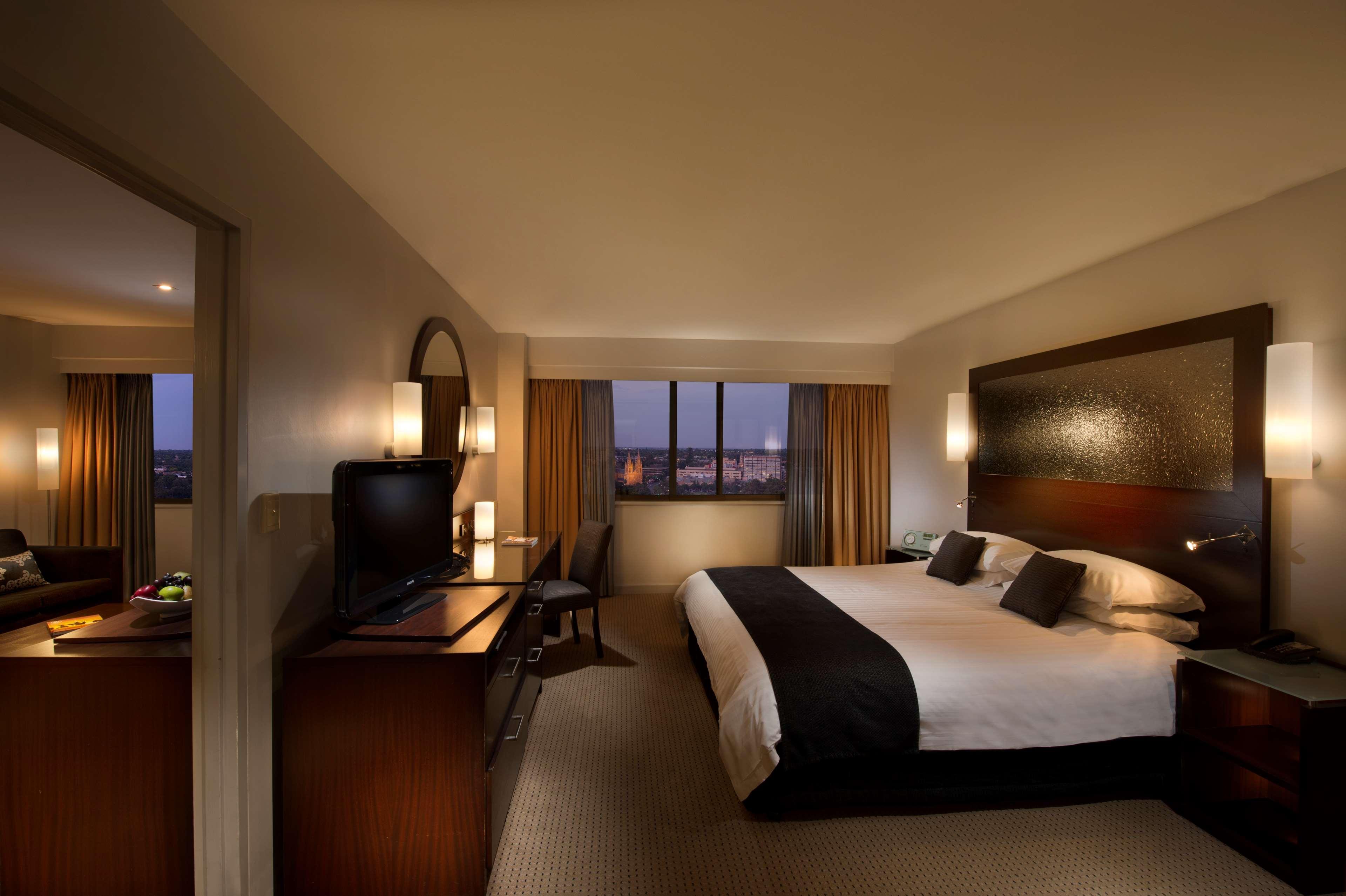 Отель Stamford Plaza Аделаида Экстерьер фото The photo features a modern hotel room that includes a large bed with dark and light bedding, complemented by two pillows. The room is well-lit with warm lighting and has a contemporary aesthetic. To one side, there is a wooden dresser with a flat-sc