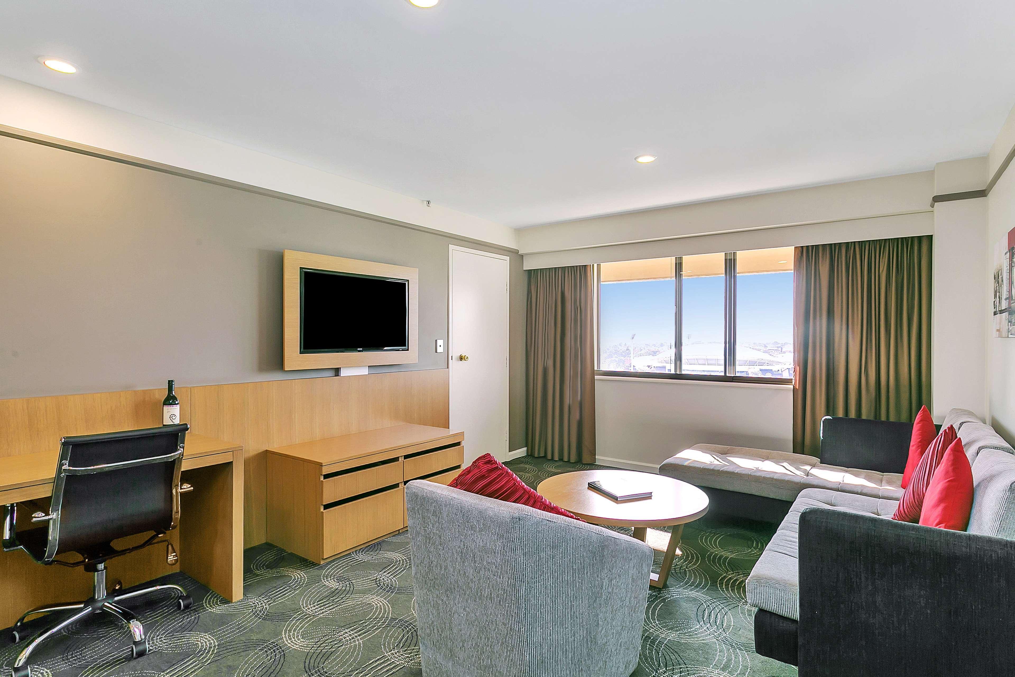 Отель Stamford Plaza Аделаида Экстерьер фото The photo shows a modern hotel room or suite that features a cozy seating area. There is a comfortable sofa with red accent pillows, a round coffee table in the center, and a sleek desk with an office chair. On the wall, there is a flat-screen TV mou