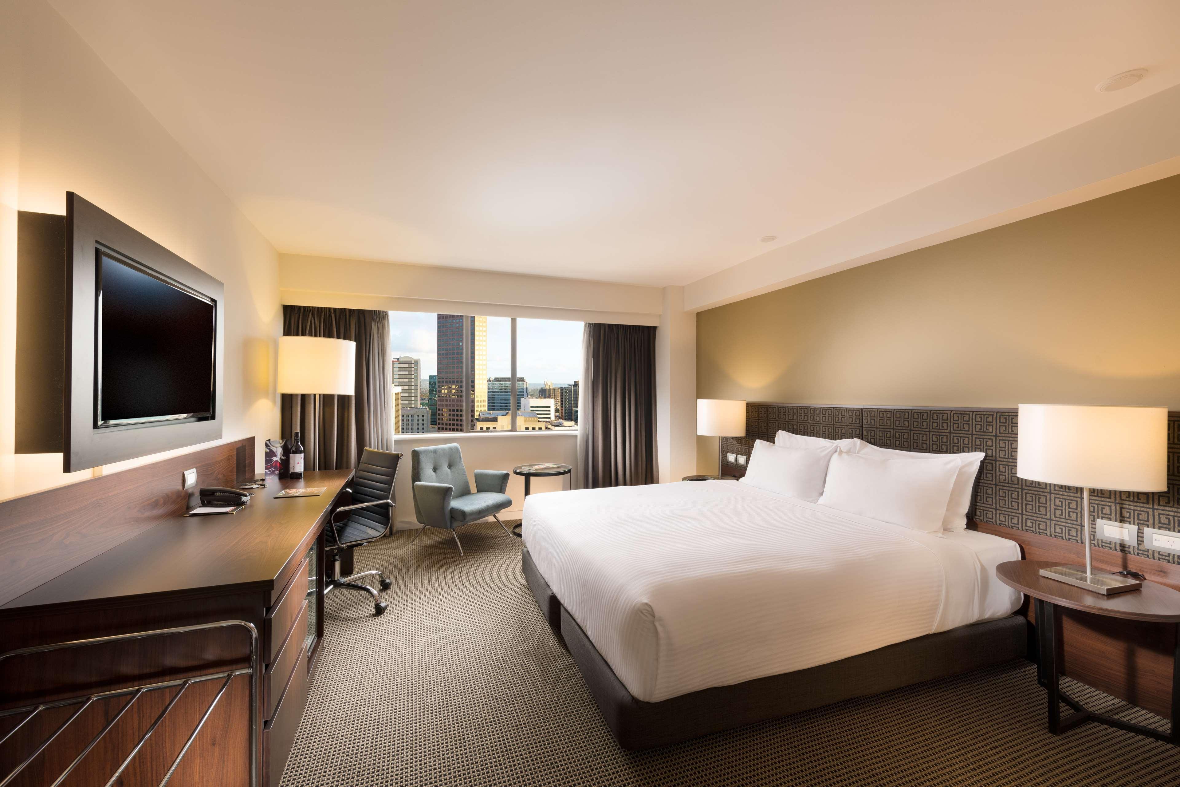 Отель Stamford Plaza Аделаида Экстерьер фото The photo shows a modern hotel room with a contemporary design. It features a large bed with white linens, a bedside table on either side with lamps, and a dark wood accent wall. There's a work desk with an office chair and a flat-screen television m