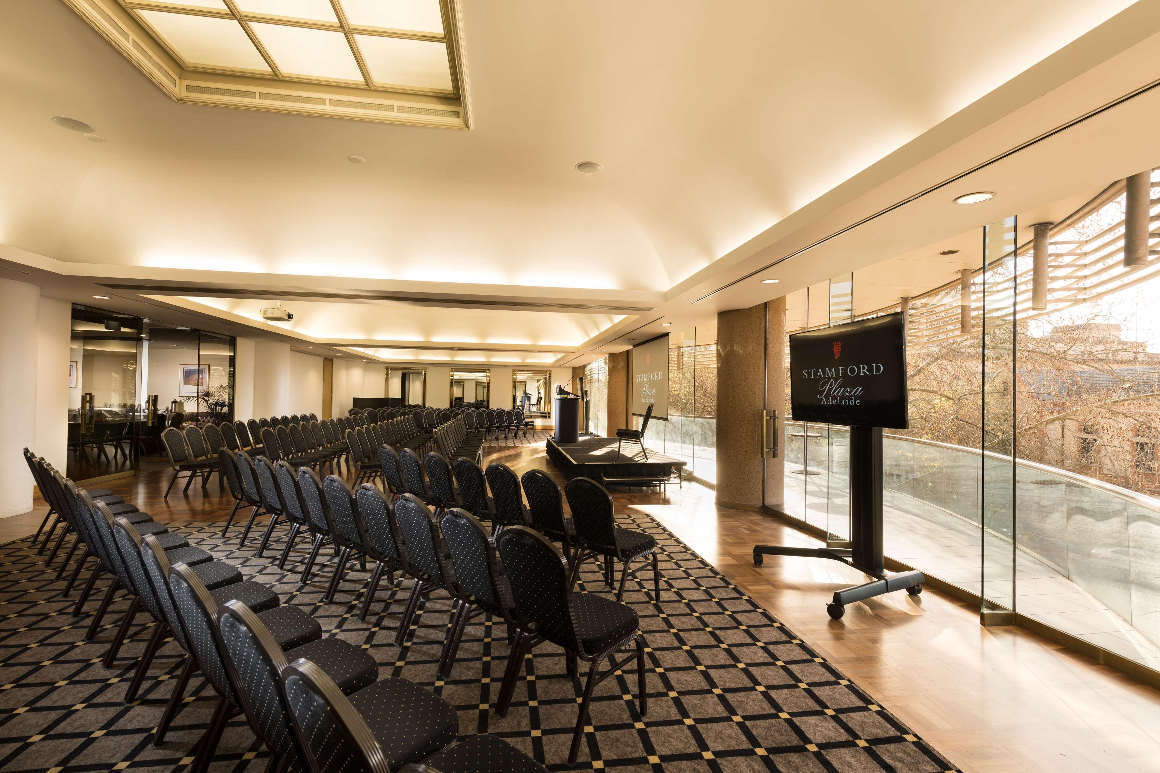 Отель Stamford Plaza Аделаида Экстерьер фото The photo shows a large, elegantly designed event space or meeting room. There are rows of black chairs set up facing a presentation area. The flooring features a patterned carpet, and large windows allow natural light to enter the room. On one side,