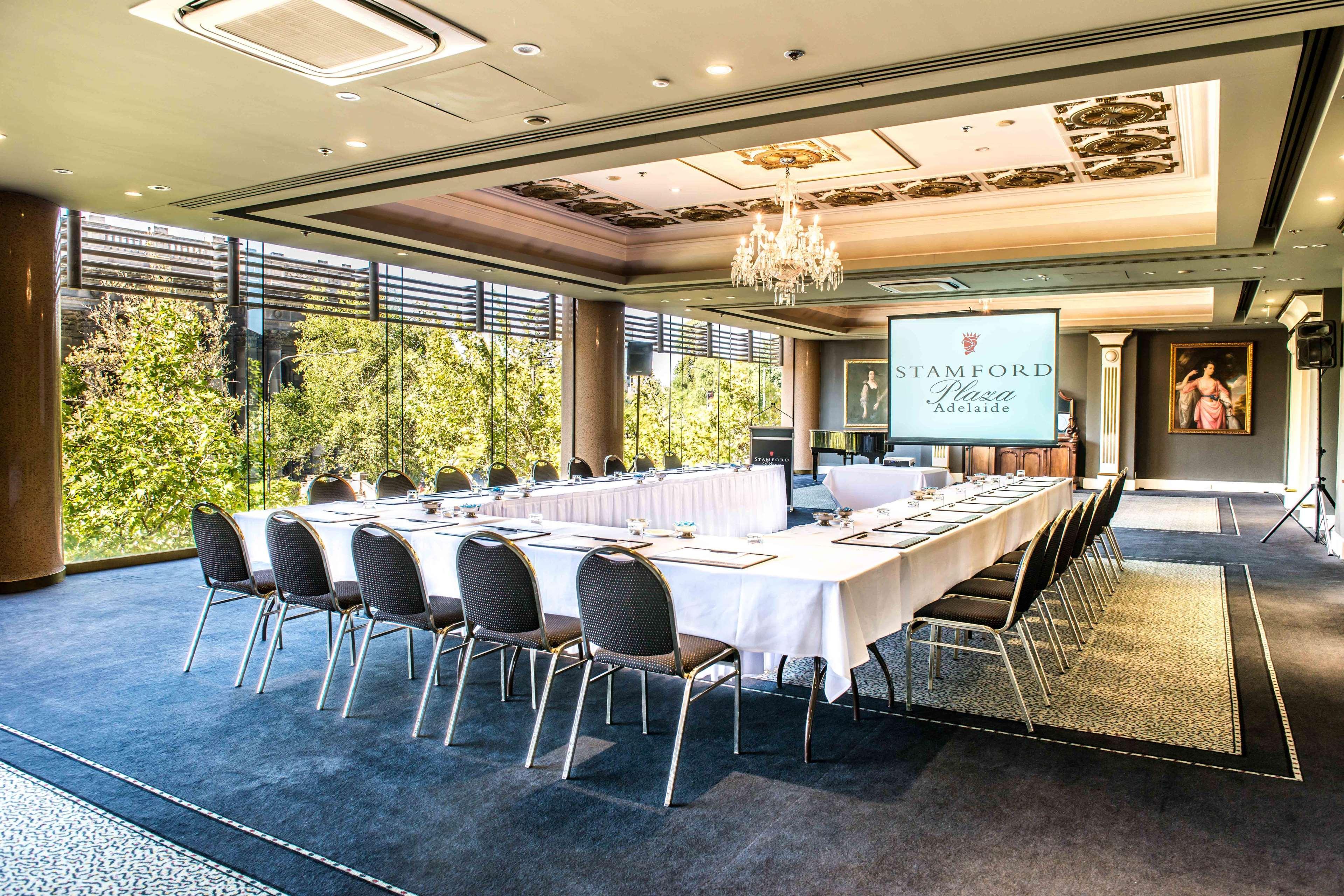 Отель Stamford Plaza Аделаида Экстерьер фото The photo shows a stylish conference room set up for a meeting or event. There is a long rectangular table covered with a white tablecloth, surrounded by black chairs. A large screen displays the name "Stamford" along with a logo. The room features l
