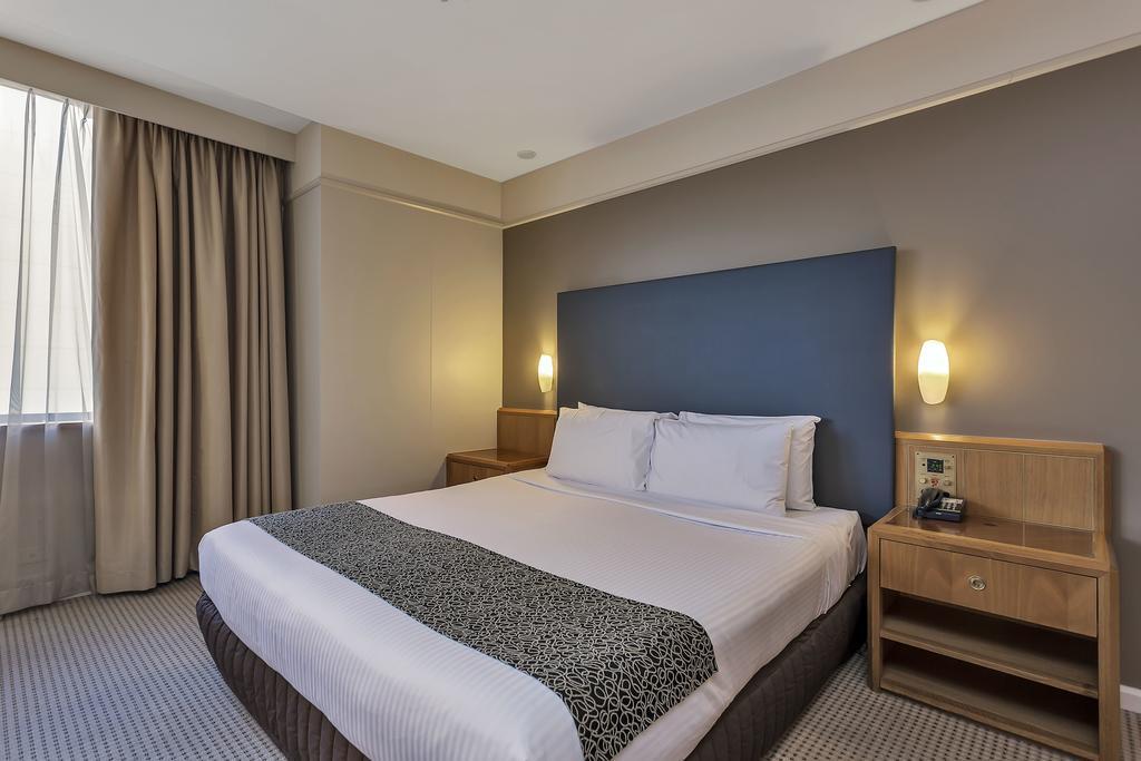 Отель Stamford Plaza Аделаида Экстерьер фото The photo shows a modern hotel room. It features a double bed with a white duvet and a decorative grey and black throw. The headboard is a solid dark color. There are two bedside tables on either side of the bed, each with a lamp. Natural light enter